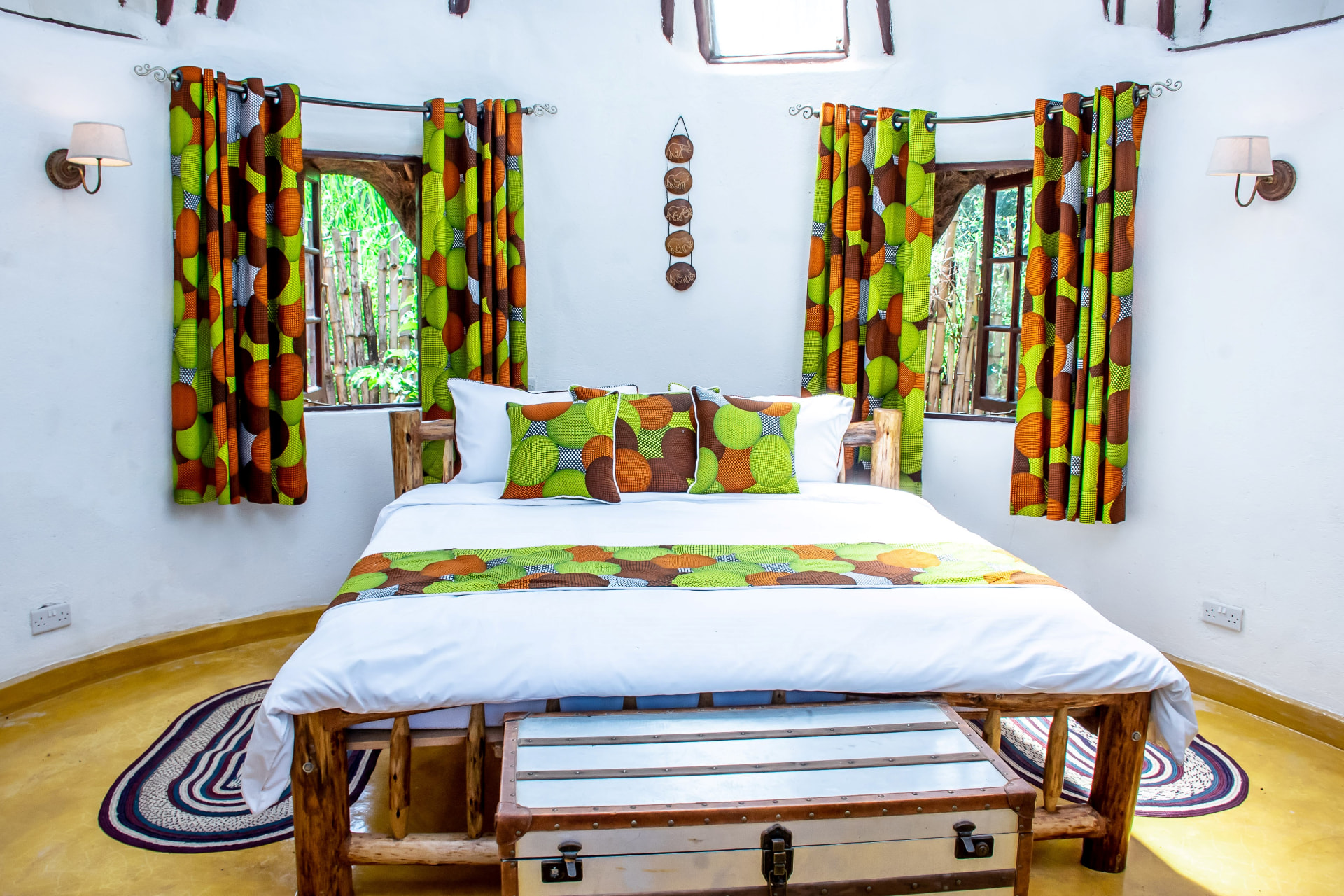 Round cottage bedroom with bold green and orange Ankara print curtains, matching cushions and a timber bed on a polished ochre floor