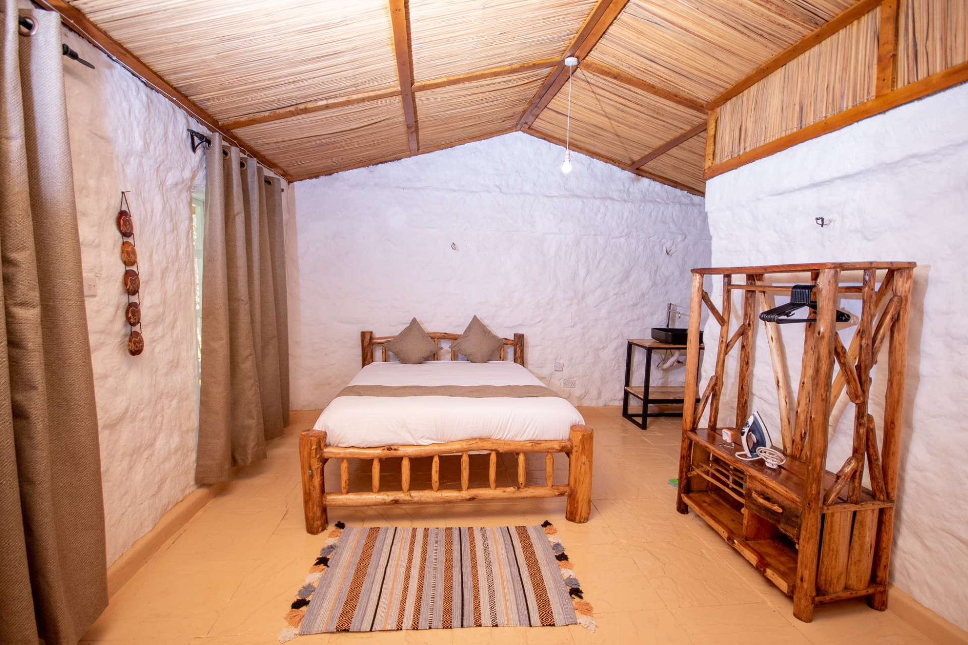 Rustic guest suite with reed and timber ceiling, lime-washed stone walls, hand-built log bed and woven striped rug