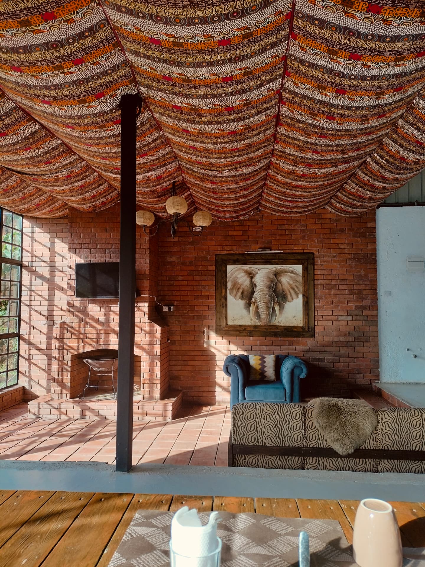 Lodge lounge with draped African print fabric canopy, exposed brick fireplace wall, blue velvet armchair and animal-print daybed