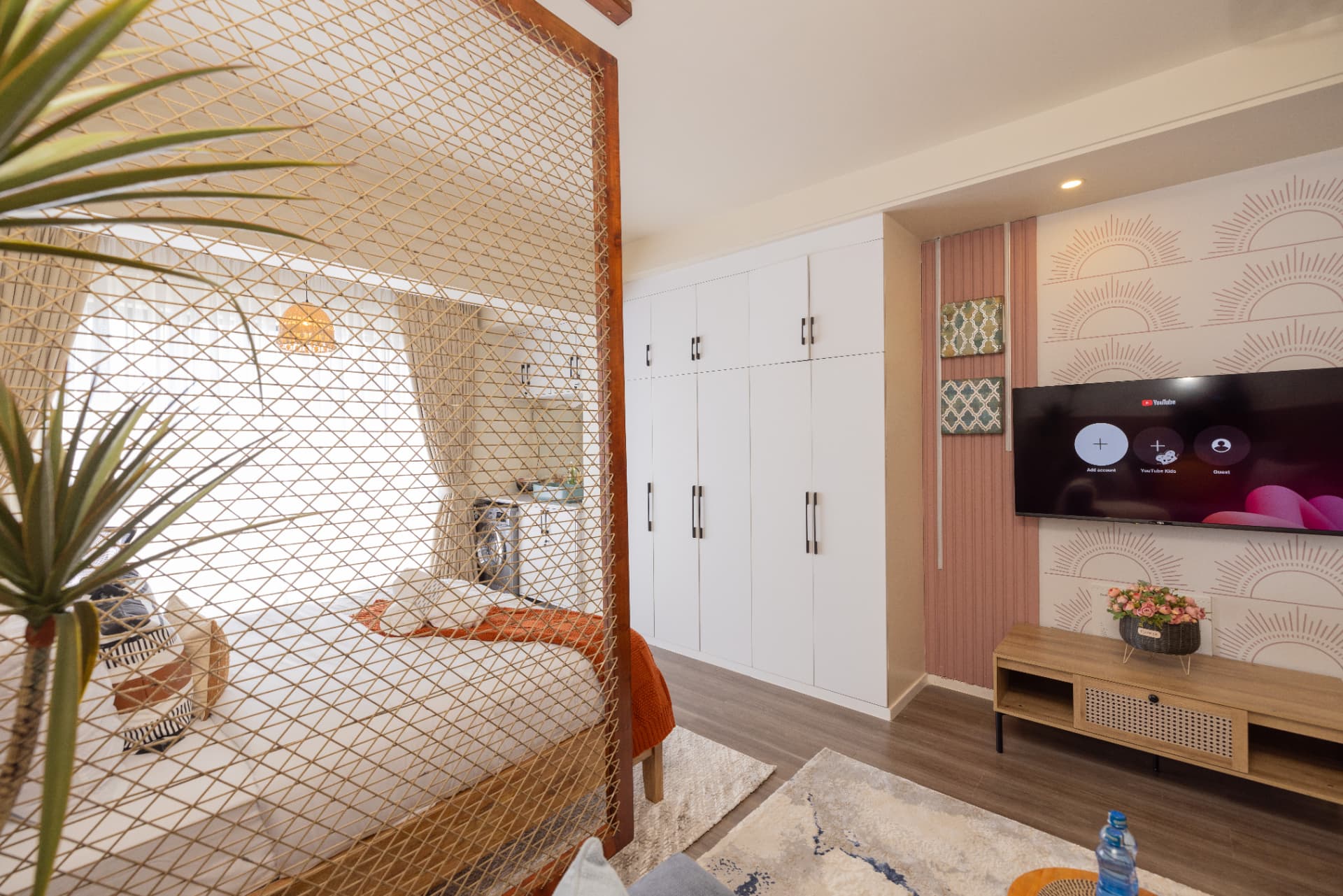 Studio apartment living area with sunburst patterned wallpaper, fluted blush panelling, wall-mounted television and rattan media unit
