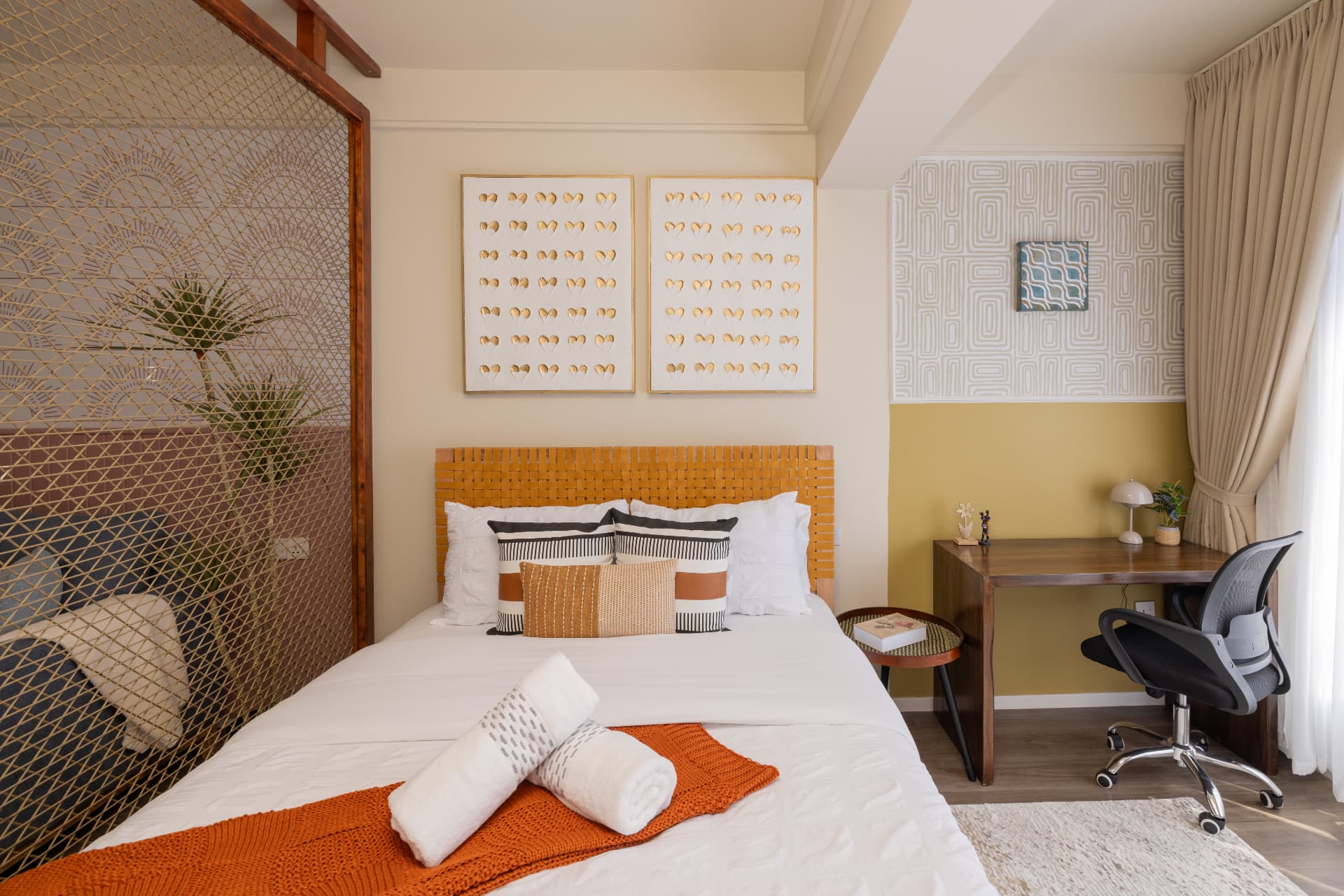 Studio apartment bedroom with woven leather headboard, ochre accent wall, timber desk and hand-woven screen dividing the space